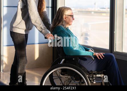 Tochter, die ihrer älteren Mutter im Rollstuhl hilft, während sie am Flughafen auf ihren Flug wartet Stockfoto