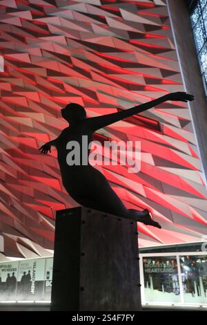 Belfast, Nordirland - 22. Dezember 2024 - Silhouette der öffentlichen Kunstskulptur „Titanica“ vor der Titanic Belfast mit roten Lichtern im Hintergrund. Außenansicht des Titanic Belfast Museumsgebäudes, beleuchtet nachts während der Weihnachtsfeiertage im Titanic Quarter in Belfast City. Stockfoto