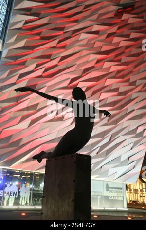 Belfast, Nordirland - 22. Dezember 2024 - Silhouette der öffentlichen Kunstskulptur „Titanica“ vor der Titanic Belfast mit roten Lichtern im Hintergrund. Außenansicht des Titanic Belfast Museumsgebäudes, beleuchtet nachts während der Weihnachtsfeiertage im Titanic Quarter in Belfast City. Stockfoto