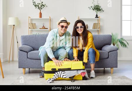 Junge positive Paare packen ihren Koffer, bereiten sich auf die Urlaubsreise vor und schauen in die Kamera. Stockfoto