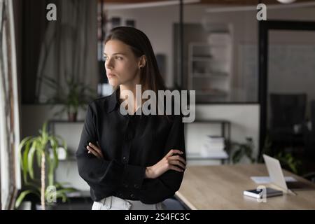 Junge Frau, die neben dem Fenster steht und mit kontemplativem Ausdruck nach draußen blickt Stockfoto
