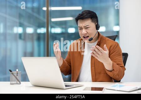 Wütender Mann mit Headset, der im Büro auf einem Laptop sitzt, unzufriedener Mitarbeiter, der den Gesprächspartner anschreit, nicht in der Lage ist, mit dem Kunden zu kommunizieren. Stockfoto
