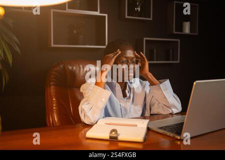 Junge Geschäftsfrau, die schwere Kopfschmerzen hat, während sie spät in der Nacht an einem Laptop arbeitet, Schläfe berührt, unter Migräne leidet aufgrund von Stress und Überlastung, am Schreibtisch im dunklen Büro sitzt Stockfoto