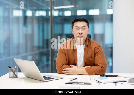 Porträt eines selbstbewussten seriösen Geschäftsmannes, eines asiatischen Mannes, der in die Kamera schaut, eines Mannes hinter Papierkram und eines Laptops, der in die Kamera schaut. Stockfoto