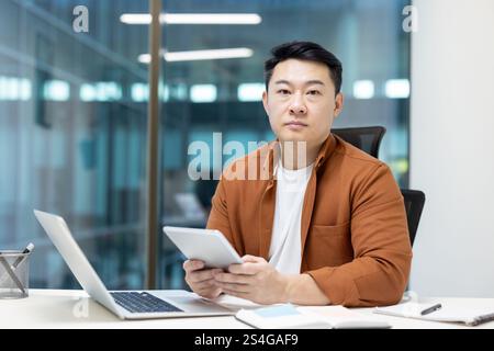 Porträt eines ernsten und konzentrierten Mannes am Arbeitsplatz im Büro. Asiatischer Mann, der in die Kamera schaut, während er einen Tablet-Computer hält. Erfahrener Entwickler von Programmierern für Büromitarbeiter. Stockfoto