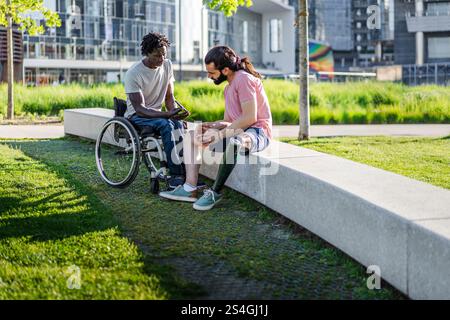 Zwei Freunde, einer mit einer Beinprothese und der andere mit einem Rollstuhl, sitzen auf einer Betonbank in einem modernen Stadtpark. Engagierte sich für ein bedeutendes c Stockfoto