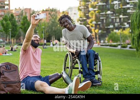 Zwei Freunde, einer mit einer Beinprothese und der andere mit einem Rollstuhl, lächeln während eines Selfie in einem grünen Stadtpark. Das Bild betont einschl Stockfoto