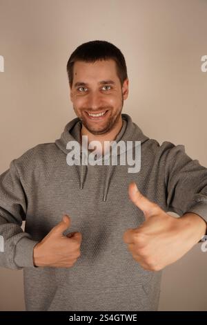 Ein fröhlicher Mann zeigt mit beiden Händen einen Daumen hoch und strahlt positive Energie aus. Er ist ein Brünette europäischer Herkunft und trägt einen grauen Hoodie. Sein lässiges Stockfoto