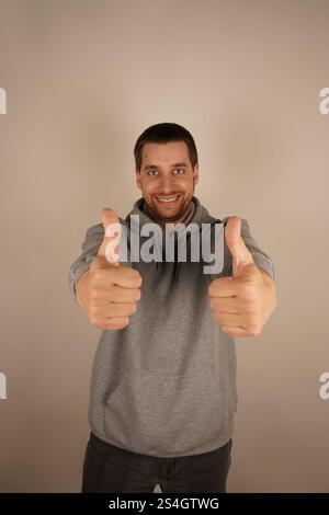 Er ist ein Brünette europäischer Herkunft und trägt einen grauen Hoodie. Sein lässiger Stil lässt ihn entspannt und ansprechbar aussehen. Ein fröhlicher Mann zeigt einen Daumen nach oben Stockfoto