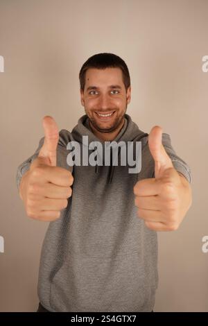 Ein fröhlicher Mann zeigt mit beiden Händen einen Daumen hoch und strahlt positive Energie aus. Er ist ein Brünette europäischer Herkunft und trägt einen grauen Hoodie. Sein lässiges Stockfoto