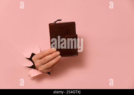 Eine Frauenhand hält eine Handtasche durch ein Loch, das in den rosa Hintergrund des Studios gerissen ist. Stockfoto