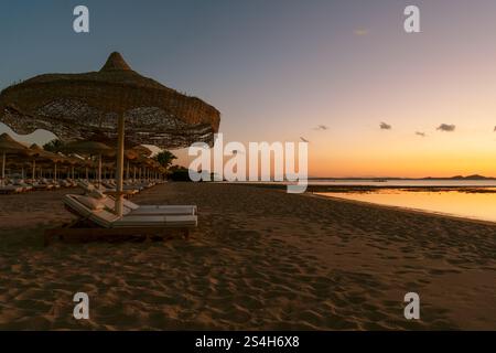 An einem Strand mit feinem Sand, Liegestühle und Sonnenschirme sitzen unter einem orangen Sonnenaufgang über dem Meer. Hochauflösendes Foto. Stockfoto