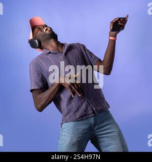 Ein fröhlicher afrikanischer Mann, der Kopfhörer trägt und ein Smartphone in der Hand hält, tanzt in der Musik energisch auf einem leuchtend blauen Hintergrund. Erfasst ein Auto Stockfoto