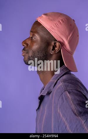 Ein Seitenprofil eines jungen Afrikaners mit rosa Mütze und lässigem Hemd, vor einem leuchtend violetten Hintergrund. Die Komposition betont Stil Stockfoto
