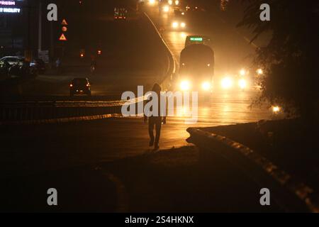 Eine einsame Figur geht die schwach beleuchtete Autobahn hinunter, umgeben von den Scheinwerfern und dem ruhigen Summen der Nacht Stockfoto