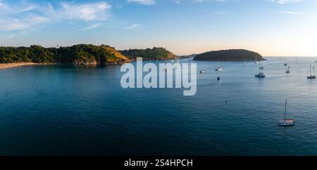 Blick aus der Vogelperspektive auf den Naiharn Beach in Phuket, Thailand, mit einer ruhigen Bucht mit verankerten Segelbooten, üppigen grünen Hügeln und einer mit Bäumen bewachsenen Insel. Stockfoto