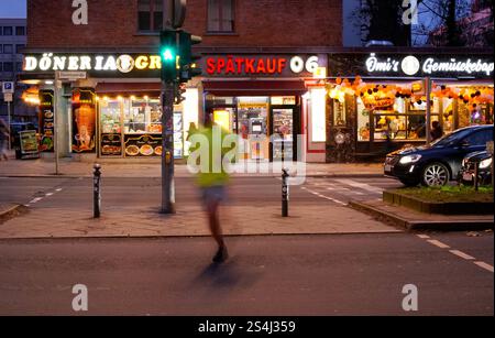 Berlin, Deutschland. Januar 2025. 11.01.2025, Berlin. Eine Person joggt an einem Wintertag in der Abenddämmerung in Berlin Schöneberg und läuft über eine Kreuzung (Aufnahme mit längerer Belichtungszeit). Hinter ihm sehen Sie die Anzeigen eines Drogengeschäfts, eines Lebensmittelläden und einer Snackbar für Gemüsekebabs. In Berlin haben viele Migranten ihre eigenen Doener- oder Dönerläden eingerichtet. Kredit: Wolfram Steinberg/dpa Kredit: Wolfram Steinberg/dpa/Alamy Live News Stockfoto