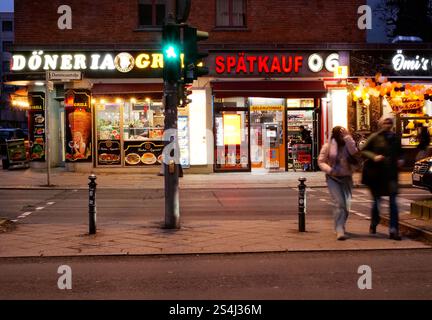 Berlin, Deutschland. Januar 2025. 11.01.2025, Berlin. Menschen gehen an einem Wintertag in der Abenddämmerung in Berlin Schöneberg über eine Kreuzung (Aufnahme mit längerer Belichtungszeit). Hinter ihnen sehen Sie die Leuchtwerbung einer Drogerie, eines 24-Stunden-Geschäfts und einer Snackbar für Gemüsekebabs. In Berlin haben viele Migranten ihre eigenen Doener- oder Dönerläden eingerichtet. Kredit: Wolfram Steinberg/dpa Kredit: Wolfram Steinberg/dpa/Alamy Live News Stockfoto