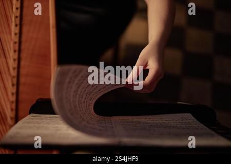 Draufsicht auf die Hand der Harfenspielerin, die Notenblätter mit Notizen umschlägt, während die Musik im Raum mit dezentem Licht und Kopierraum gespielt wird Stockfoto