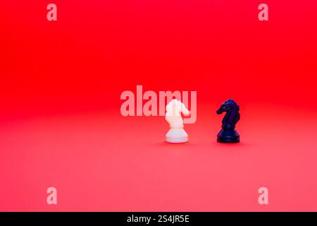 Ein weißes und ein schwarzes Ritterschach stehen einander gegenüber vor einem auffälligen roten Hintergrund, was das Spiel von Strategie und Wettbewerb unterstreicht Stockfoto