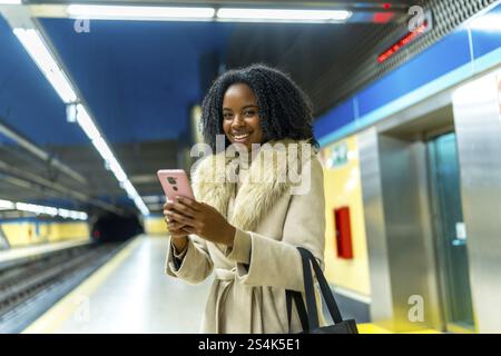 Lächelnde Geschäftsfrau, die ihr Mobiltelefon benutzt, während sie am Bahnhof auf die U-Bahn wartet, verkörpert modernen urbanen Lebensstil und Konnektivität Stockfoto