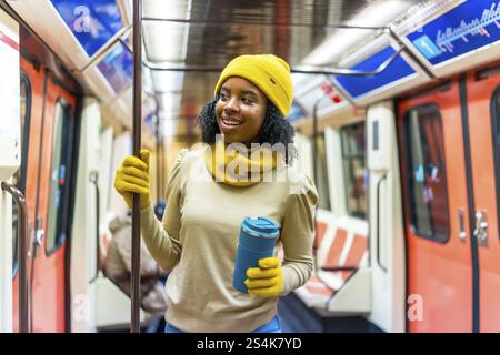Lächelnde Frau mit gelbem Wollmütze und Handschuhen, die einen wiederverwendbaren Becher in der Hand halten, während sie in einem U-Bahn-Auto steht Stockfoto
