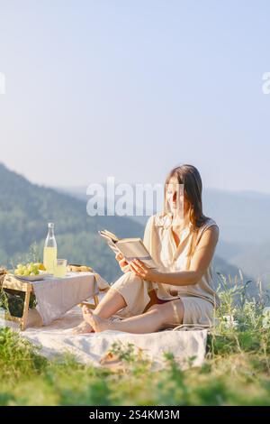 Frau genießt ein friedliches Picknick in den Bergen, liest ein Buch und schlürft Limonade an einem sonnigen Tag. Stockfoto