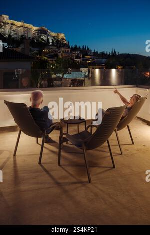 Ein Mann und eine Frau sitzen mit einer Flasche Wein in der Abenddämmerung in Liegestühlen auf einer Dachterasse in Athen und schauen auf die beleuchtet Stockfoto