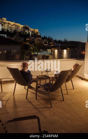 Ein Mann und eine Frau sitzen mit einer Flasche Wein in der Abenddämmerung in Liegestühlen auf einer Dachterasse in Athen und schauen auf die beleuchtet Stockfoto