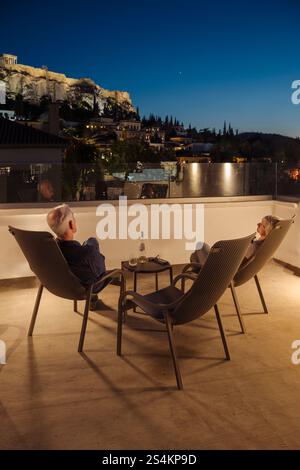 Ein Mann und eine Frau sitzen mit einer Flasche Wein in der Abenddämmerung in Liegestühlen auf einer Dachterasse in Athen und schauen auf die beleuchtet Stockfoto