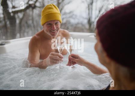 Seniorenpaar baden im Wannen-Whirlpool während des kalten Wintertages und toasten mit Champagner. Romantischer Wellness-Kurzurlaub mit Spa am Valentinstag-Wochenende. Stockfoto