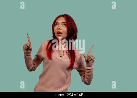 Studio-Aufnahme einer überraschten jungen Frau mit rotem Haar, die mit Zeigefingern auf türkisfarbenem Hintergrund nach oben zeigt Stockfoto