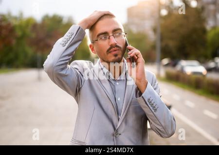 Porträt eines verzweifelten Geschäftsmannes, der am Telefon spricht. Junger Geschäftsmann, der auf der Straße steht und ein Smartphone benutzt. Stockfoto