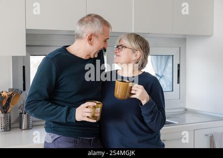 Seniorenpaare teilen einen freudigen Moment in einer modernen Küche, jedes hält eine Kaffeetasse. Ihre Wärme und Verbundenheit schaffen eine Atmosphäre des Komforts und der Co Stockfoto