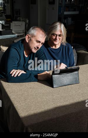 Ein älteres Ehepaar sitzt an einem Tisch in einem gemütlichen Wohnzimmer, mit einem digitalen Tablet beschäftigt und scheint die gemeinsame Freizeit zu genießen, um eine Mutter zu teilen Stockfoto