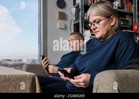 Ein Seniorenpaar sitzt in ihrem gemütlichen Wohnzimmer, jedes mit einem Smartphone. Das Bild erfasst moderne Konnektivität und Entspannung in einem gut beleuchteten Spa Stockfoto