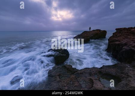 Ein dramatischer Blick auf das Meer in Cabo Cervera, Torrevieja, Alicante, Eine einsame Figur steht auf Felsen und überblickt die Wellen, die gegen die zerklüftete Küste stürzen Stockfoto
