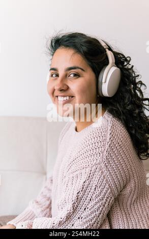 Eine junge lateinfrau sitzt auf einer gemütlichen Couch, trägt Kopfhörer und einen Strickpullover. Sie lächelt, strahlt ein Gefühl von Entspannung und Freude aus und nimmt ein Stockfoto