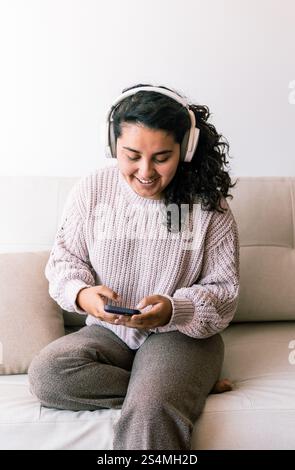 Eine junge lateinfrau sitzt bequem auf einer Couch, trägt Kopfhörer und schaut auf ihr Handy. Sie strahlt Entspannung und Glück aus und genießt ihre Zeit Stockfoto
