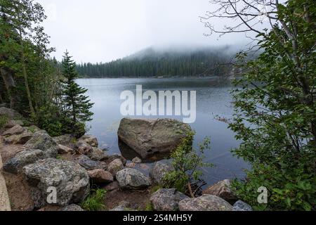 Ein ruhiger Blick auf einen ruhigen See, umgeben von üppigem Grün und felsigem Gelände unter einer Decke aus Morgennebel, gefangen in der Ruhe von Colorado's W Stockfoto