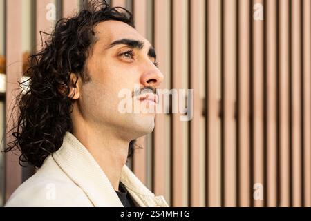 Ein stilvoller Mann mit lockigen Haaren und Schnurrbart blickt in die Ferne, gekleidet in einer beigen Jacke vor dem Hintergrund vertikaler Holzpaneele auf einem sunn Stockfoto