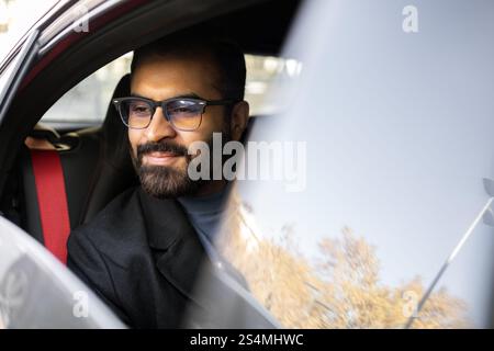 Ein Indianer, der in einem dunklen Wintermantel und einer Brille gekleidet ist, wird während der Fahrt mit seinem Auto sanft lächelnd gesehen, was ein Gefühl von Zufriedenheit und Komfort vermittelt. Stockfoto