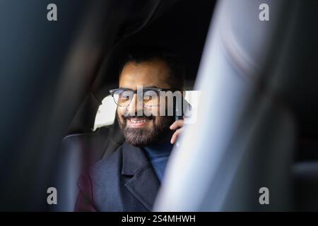 Ein lächelnder Indianer in Wintermantel und Pullover, der auf einem Handy spricht, während er im Auto sitzt und von der Kamera wegblickt. Stockfoto