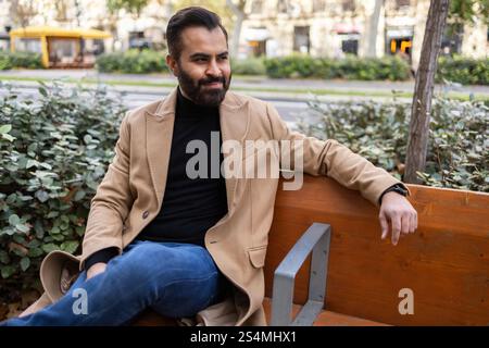 Ein Inder, der als Geschäftsmann in einem stilvollen beigefarbenen Mantel und Jeans gekleidet ist, entspannt sich auf einer Parkbank und blickt nachdenklich in üppigem Grün weg. Stockfoto