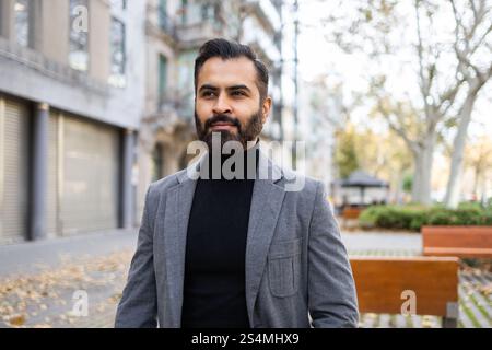 Indischer Geschäftsmann in Winterkleidung, mit stilvollem Bart, souverän auf einer Straße der Stadt stehend und wegblickend. Stockfoto