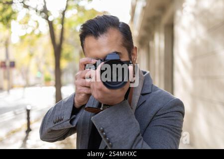 Ein Indianer, der als Fotograf mit grauem Mantel gekleidet ist, konzentriert sich darauf, mit seiner professionellen Kamera Bilder in einer grünen, sonnendurchfluteten Stadt aufzunehmen Stockfoto