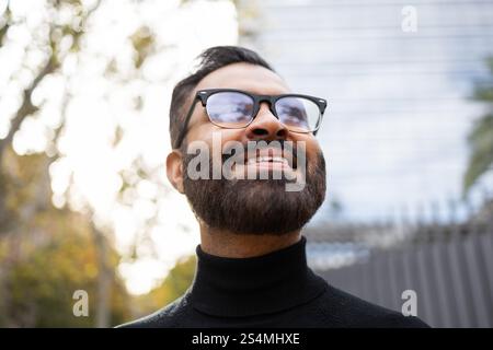 Von unten aus genießt der fröhliche indische Geschäftsmann mit Sonnenbrille und schwarzem Rollkragen einen sonnigen Wintertag im Freien und blickt von der Kamera weg Stockfoto