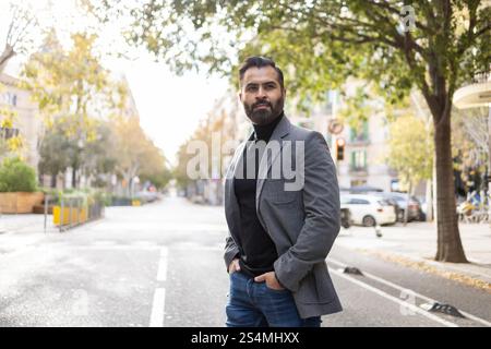 Stilvoller indischer Geschäftsmann mit gut geschnittenem Bart steht selbstbewusst auf einer von Bäumen gesäumten städtischen Straße, gekleidet in einem scharfen grauen Blazer und dunklem T-Shirt Stockfoto