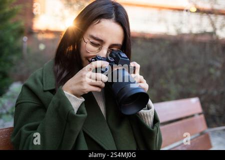 Eine Frau mit grüner Jacke sitzt auf einer Parkbank und lächelt, während sie Fotos mit einer professionellen Kamera macht Stockfoto
