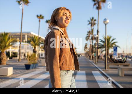 Eine Frau mit gemischter Rasse in lässiger Jacke schlendert entlang einer sonnigen Promenade, die von Palmen gesäumt ist. Die warme Beleuchtung und der klare Himmel sorgen für eine entspannte und fröhliche Atmosphäre Stockfoto